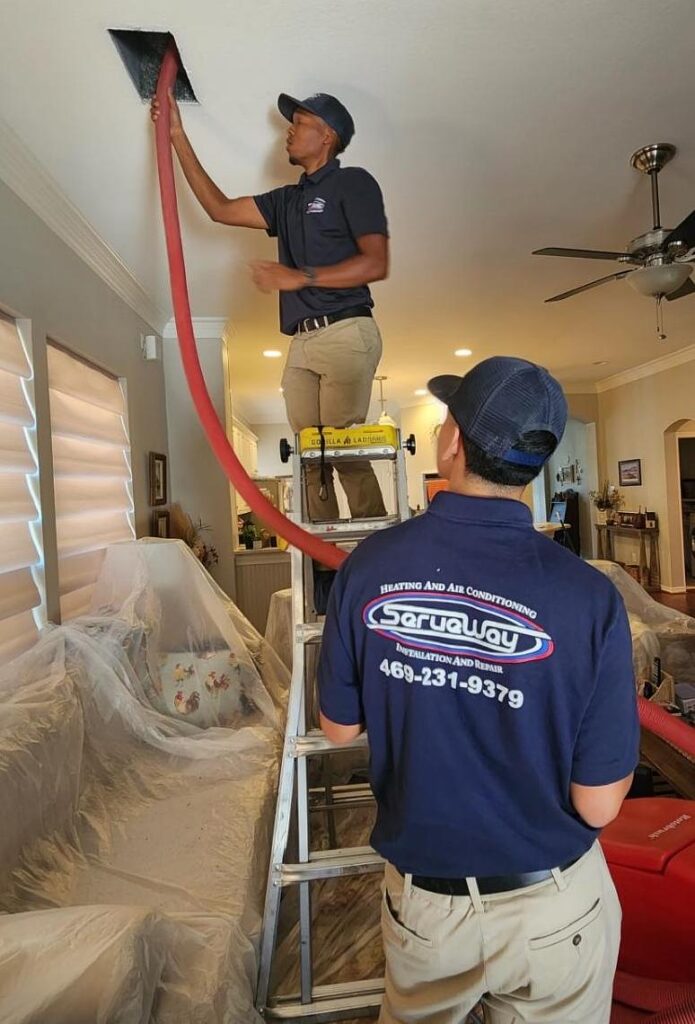 Two technicians in navy uniforms perform ceiling duct cleaning in a living room.